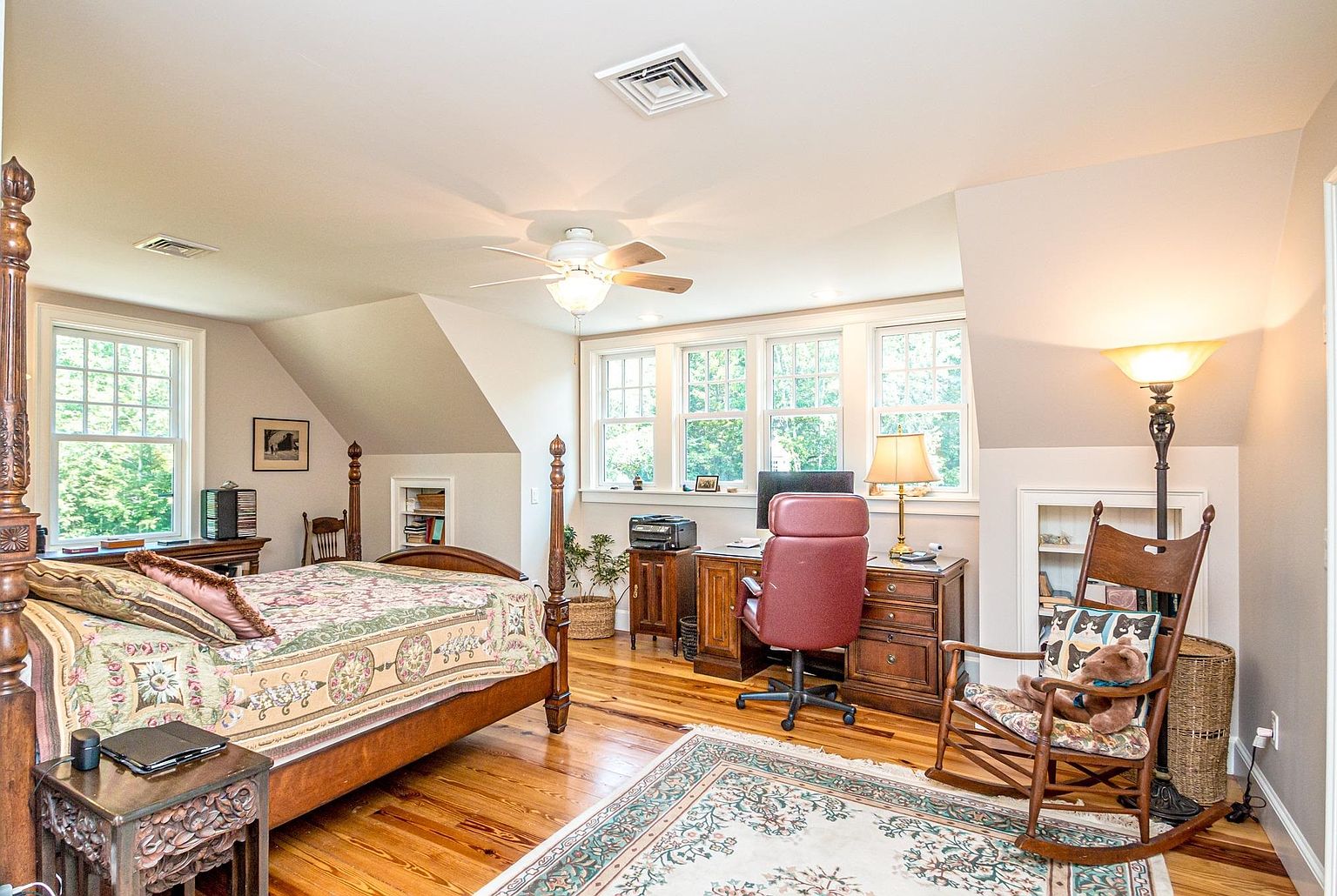 This cozy, multi-functional bedroom features a four-poster wooden bed dressed in a patterned quilt and a dedicated workspace with a desk and office chair. The room is illuminated by a ceiling fan and a floor lamp, with natural light streaming in through a row of windows. A wooden rocking chair with a teddy bear sits in the corner, adding a charming, lived-in feel to the space.