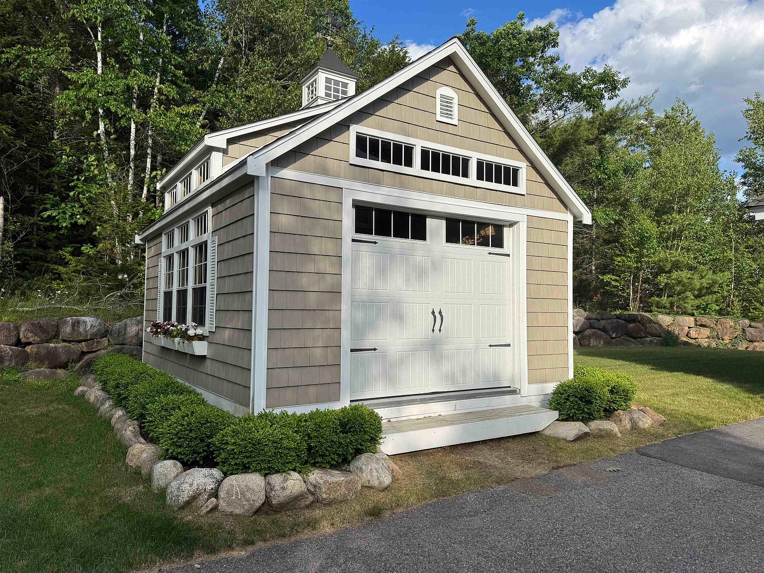 This charming, detached garage or shed features light-colored shingle siding, white trim, and a classic cupola atop its gabled roof. The structure includes a white carriage-style garage door with transom windows, a row of clerestory windows, and a side window box filled with flowers. The building is neatly landscaped with manicured shrubs and a stone border, set against a backdrop of lush green trees.