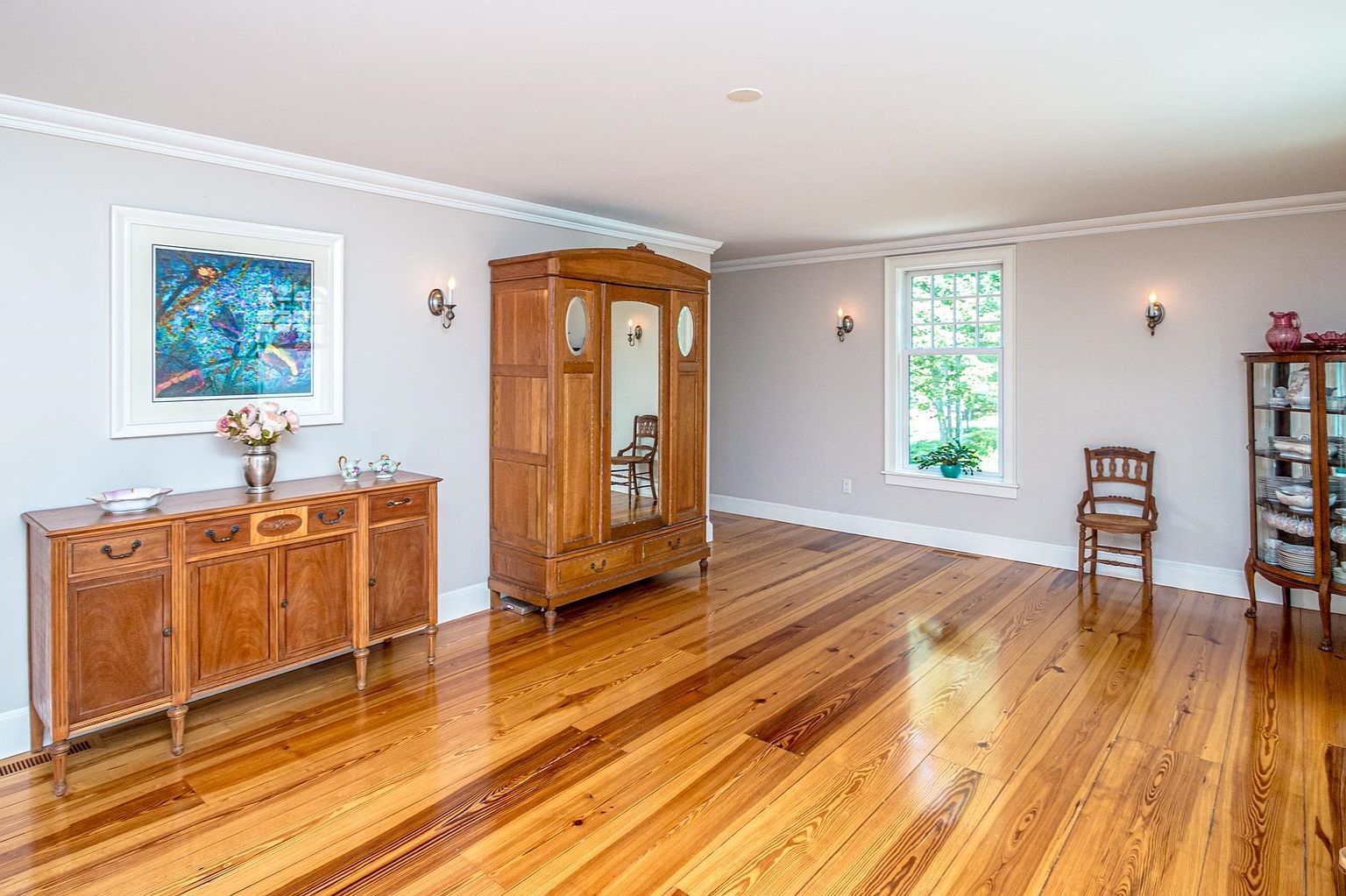 This spacious dining room features warm-toned wood flooring and light-colored walls, creating a bright and inviting atmosphere. The room is furnished with a traditional wooden sideboard, a large antique-style armoire with a mirrored center panel, and a glass-fronted china cabinet. A single wooden chair sits near a window that allows natural light to fill the space, while wall-mounted sconces provide additional ambient lighting.