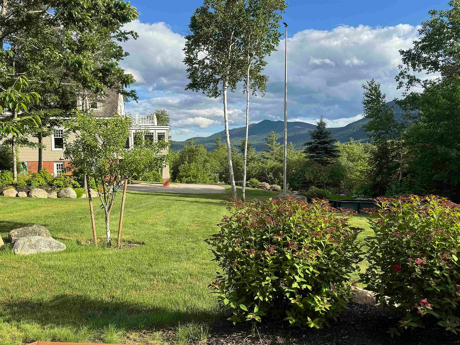 This expansive backyard view showcases a lush, well-manicured lawn framed by mature trees and vibrant flowering shrubs in the foreground. To the left, a portion of a multi-story home with a deck is visible, while the background features a stunning, panoramic vista of rolling mountains under a bright, cloudy sky. A tall flagpole stands prominently in the center, adding a classic touch to this serene and picturesque outdoor setting.