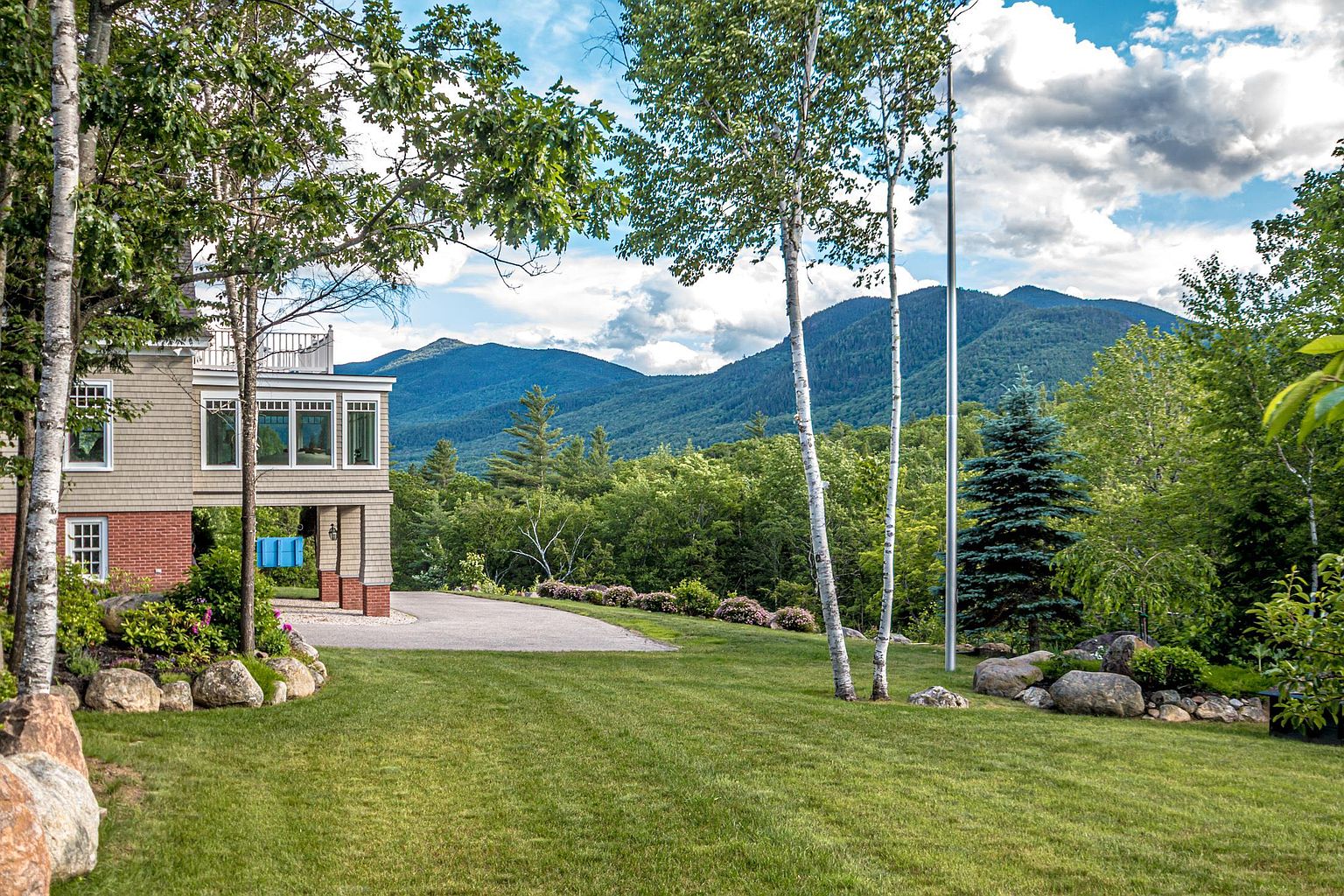 This side view of a multi-story home showcases a blend of shingle siding and brick accents, featuring a covered area with a blue porch swing. The property is set against a backdrop of lush green mountains and a clear sky with scattered clouds. A well-manicured lawn, mature birch trees, and decorative landscaping with large boulders frame the scene, creating a serene and picturesque outdoor environment.