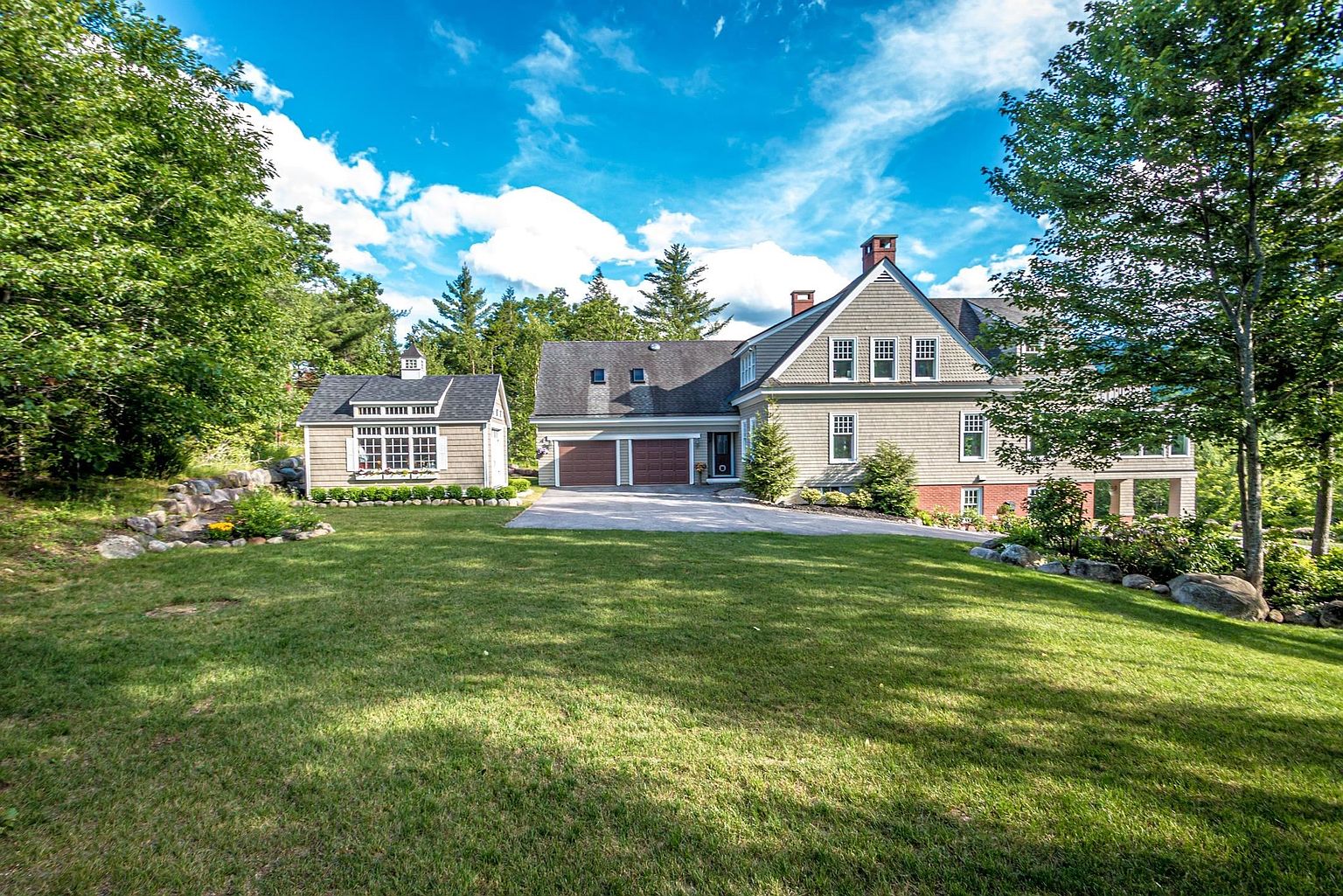 This expansive front exterior view showcases a traditional shingled home with a multi-gabled roofline and a detached garage structure. A paved driveway leads to the two-car garage, while a lush, well-manicured lawn stretches across the foreground, framed by mature trees and natural stone landscaping. The scene is captured under a bright, clear blue sky with fluffy clouds, emphasizing the property's serene and well-maintained curb appeal.