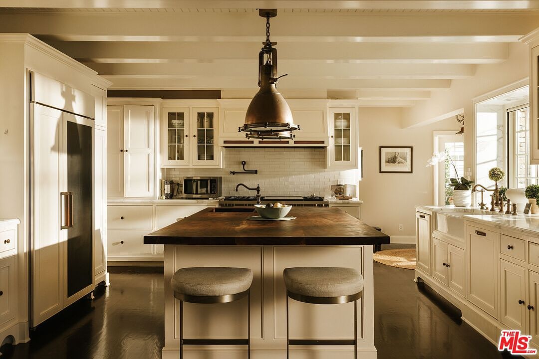 This elegant, high-end kitchen features a large central island with a dark wood countertop and two upholstered bar stools, serving as the focal point of the room. The space is defined by crisp white cabinetry, a classic subway tile backsplash, and a prominent industrial-style pendant light hanging above the island. The dark hardwood floors contrast beautifully with the bright cabinetry, creating a sophisticated and warm atmosphere perfect for culinary activities.