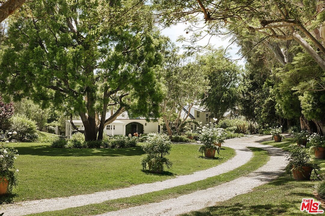 This picturesque scene captures a winding gravel driveway leading toward a charming, light-colored farmhouse nestled among lush, mature trees. The expansive, well-manicured lawn is accented by potted flowering plants and vibrant greenery, creating a serene and inviting atmosphere. The perspective is a ground-level shot that emphasizes the property's natural beauty and tranquil, park-like setting.