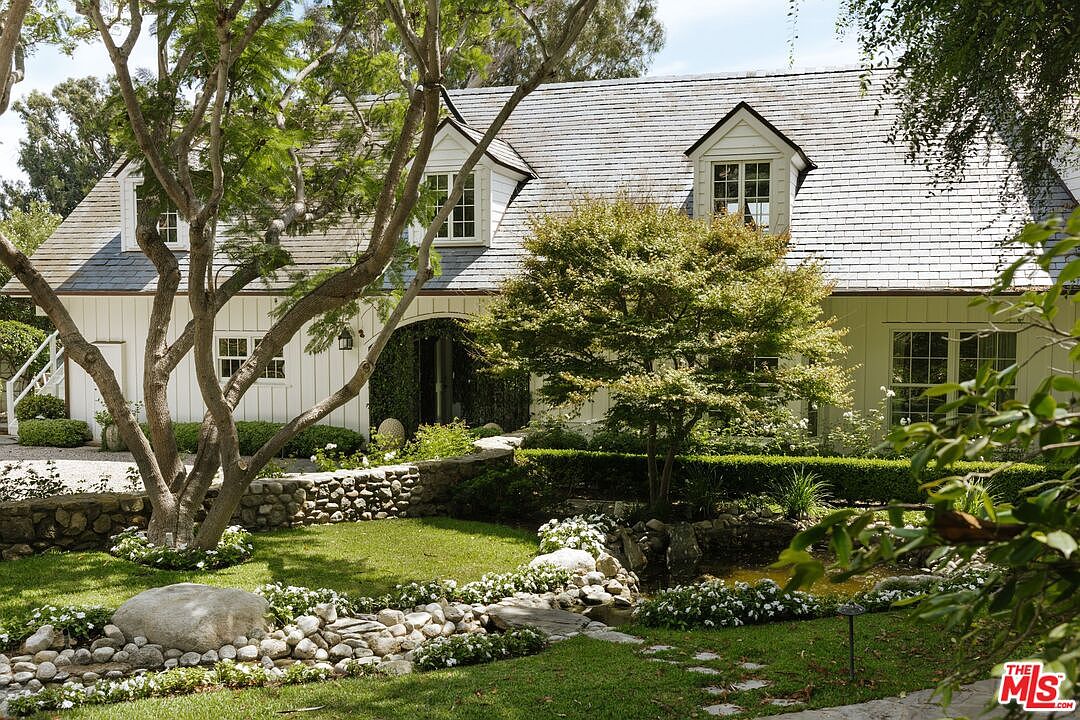 This charming, cottage-style home features white vertical siding, a steep shingled roof with dormer windows, and a stone-walled garden path leading to the entrance. The lush, meticulously landscaped front yard includes a small pond, mature trees, and vibrant greenery, creating a serene and inviting curb appeal. The perspective is a ground-level shot that captures the harmonious blend of architecture and natural beauty.