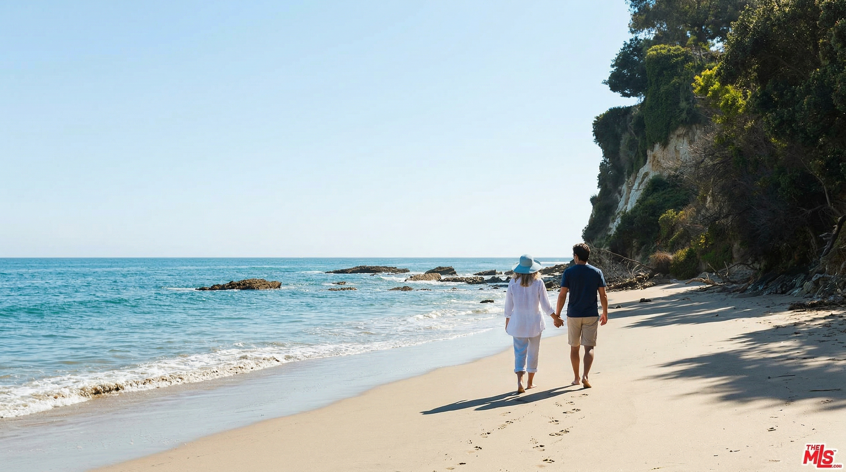 This image captures a serene, natural coastal landscape featuring a sandy beach meeting the Pacific Ocean under a clear blue sky. A rugged, tree-covered cliffside rises dramatically on the right, providing a sense of seclusion and natural beauty. The perspective is a wide-angle, eye-level shot that emphasizes the expansive horizon and the tranquil atmosphere of this private-feeling shoreline.