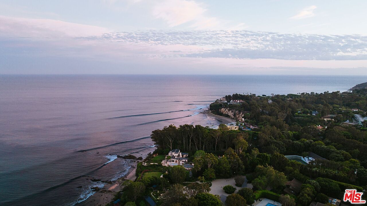 This stunning aerial view captures a luxurious coastal estate nestled along a lush, tree-lined cliffside overlooking the Pacific Ocean. The perspective highlights the expansive property grounds, the proximity to the shoreline, and the serene, natural beauty of the surrounding landscape at dusk. The scene conveys an atmosphere of exclusivity, tranquility, and high-end coastal living.