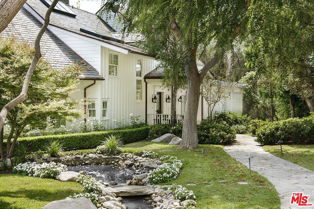 This serene garden scene features a charming stone-lined pond with a small waterfall, surrounded by lush greenery and white flowering plants. A stone pathway leads toward a beautiful white farmhouse-style home with vertical siding and a dark shingled roof, nestled among mature trees. The perspective captures a tranquil, picturesque outdoor living space that emphasizes privacy and natural beauty.