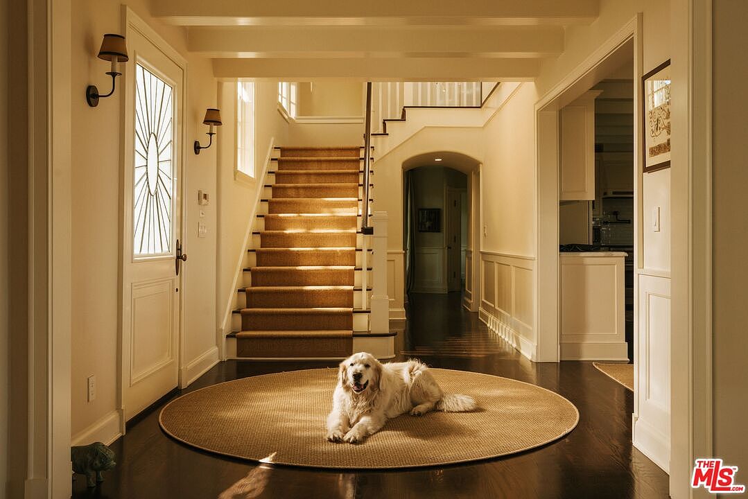 This elegant entryway features a grand staircase with a warm, carpeted runner and a classic white banister, serving as the focal point of the home. A golden retriever rests peacefully on a circular sisal rug in the center of the dark hardwood floor, creating a welcoming and serene atmosphere. The space is bathed in soft, warm lighting, highlighting the traditional architectural details and the open, inviting layout of the foyer.
