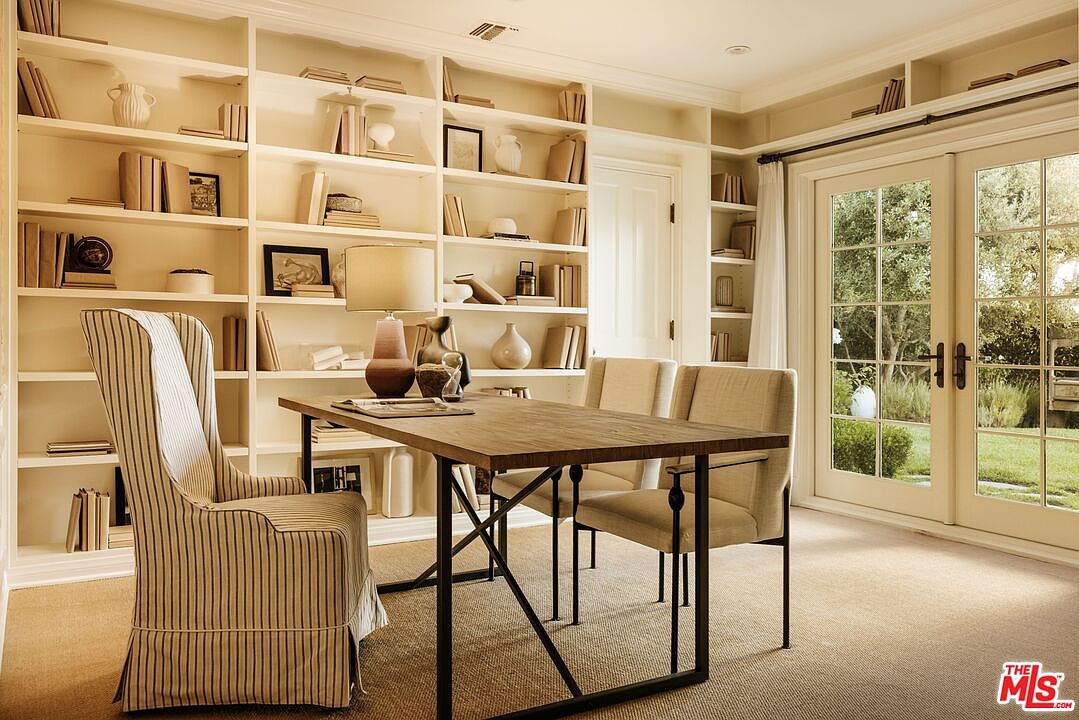 This elegant home office features floor-to-ceiling built-in bookshelves filled with books and decorative accents, creating a sophisticated and scholarly atmosphere. A rustic wooden desk with a metal frame sits at the center, accompanied by a striped upholstered armchair and two additional chairs, all set against a neutral carpet. Large glass-paned French doors allow natural light to flood the room, offering a serene view of the lush greenery outside.