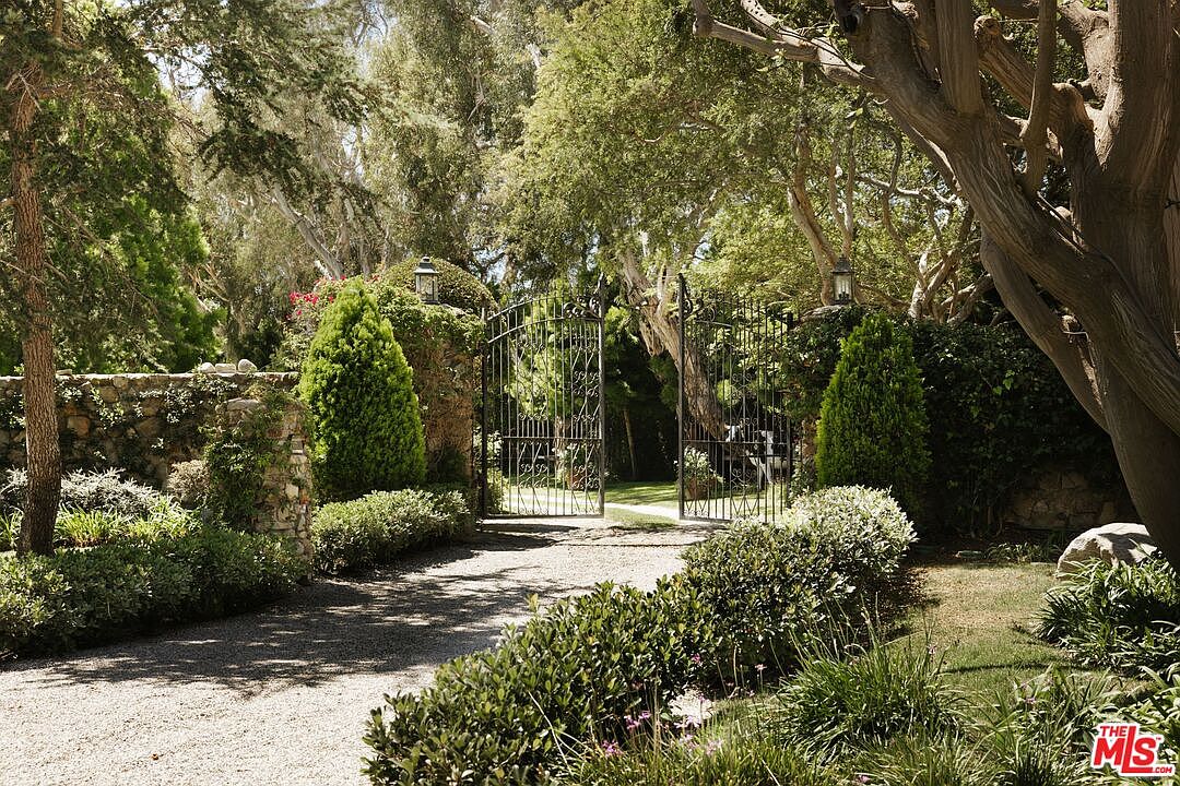 This image captures a grand, gated entrance to a private estate, framed by lush, mature trees and meticulously manicured landscaping. A gravel driveway leads through ornate black iron gates, which are flanked by stone pillars and vibrant green topiary, creating an atmosphere of exclusivity and seclusion. The perspective is eye-level, inviting the viewer to imagine passing through the gates into the serene, sun-dappled property beyond.