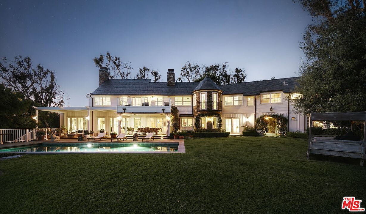 This stunning rear exterior view captures a sprawling two-story estate at dusk, featuring a classic architectural design with a prominent turret and white siding. A pristine rectangular swimming pool sits in the foreground, reflecting the warm glow of the home's interior lighting, while a manicured lawn and mature trees frame the property. The scene exudes a sense of luxury and tranquility, perfect for evening outdoor entertaining.