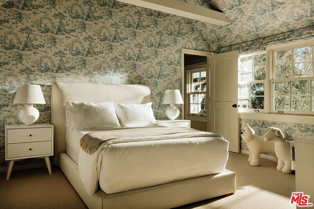 This charming bedroom features a classic toile-patterned wallpaper that extends onto the vaulted ceiling, creating a cozy and cohesive aesthetic. The room is furnished with a plush, cream-colored upholstered bed, matching nightstands with modern lamps, and two whimsical animal-shaped stools near the window. Natural light streams through the multi-pane windows, highlighting the soft textures and inviting, traditional atmosphere of the space.