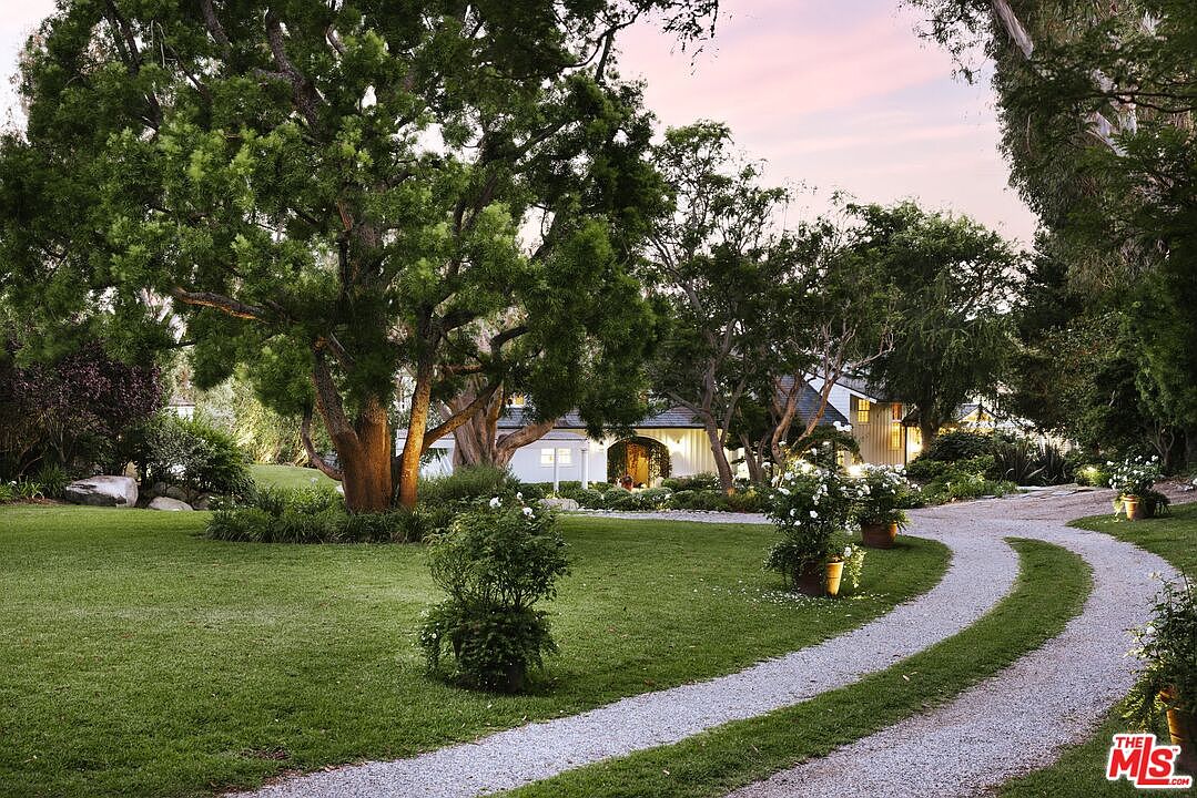 This picturesque estate features a charming, light-colored home nestled behind a lush, mature landscape and a winding gravel driveway. The scene is framed by large, leafy trees that cast soft shadows over the manicured lawn, creating a serene and inviting atmosphere. The perspective captures the home's elegant curb appeal during the golden hour, highlighting its classic architectural style and tranquil setting.