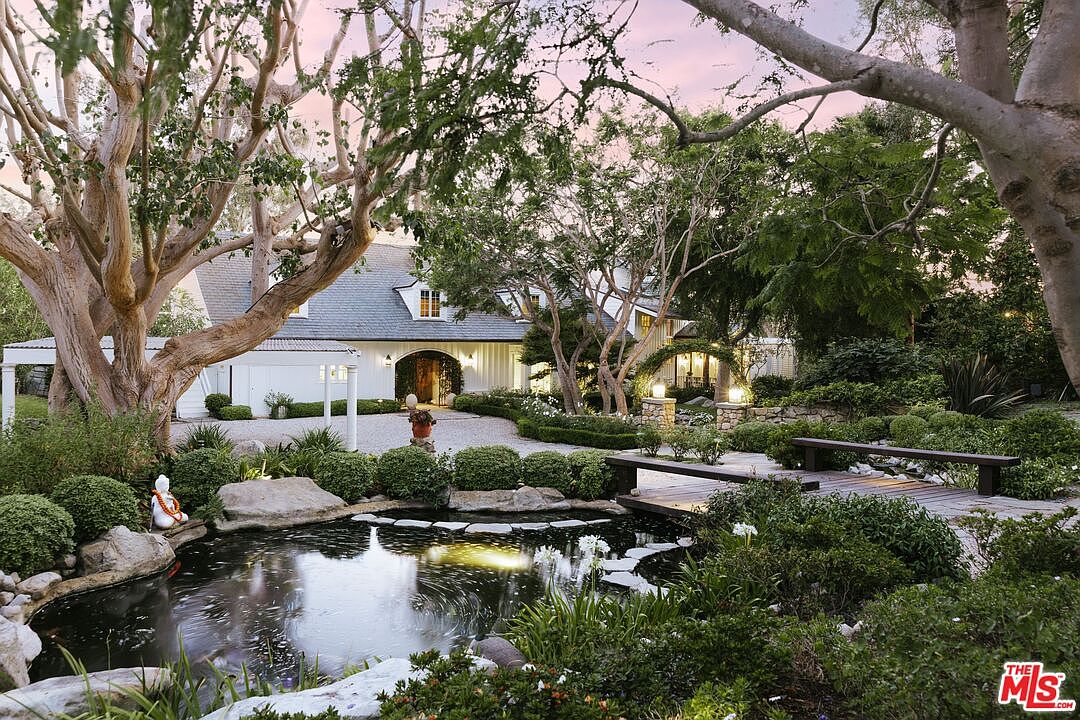 This serene estate features a beautifully landscaped front yard with a tranquil koi pond, a wooden footbridge, and lush greenery. The white farmhouse-style home is framed by mature, sprawling trees, creating a picturesque and inviting atmosphere. The perspective captures the harmonious blend of natural elements and architectural elegance, perfect for a high-end real estate presentation.