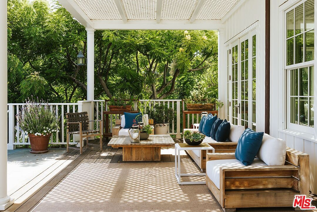 This inviting outdoor deck features a rustic wooden seating area with plush white cushions and vibrant blue throw pillows, centered around a low-profile wooden coffee table. The space is shaded by a white lattice pergola, casting a patterned shadow across the deck, and is framed by a white railing that overlooks lush green trees. The overall aesthetic is a serene, coastal-inspired retreat perfect for relaxation and entertaining.