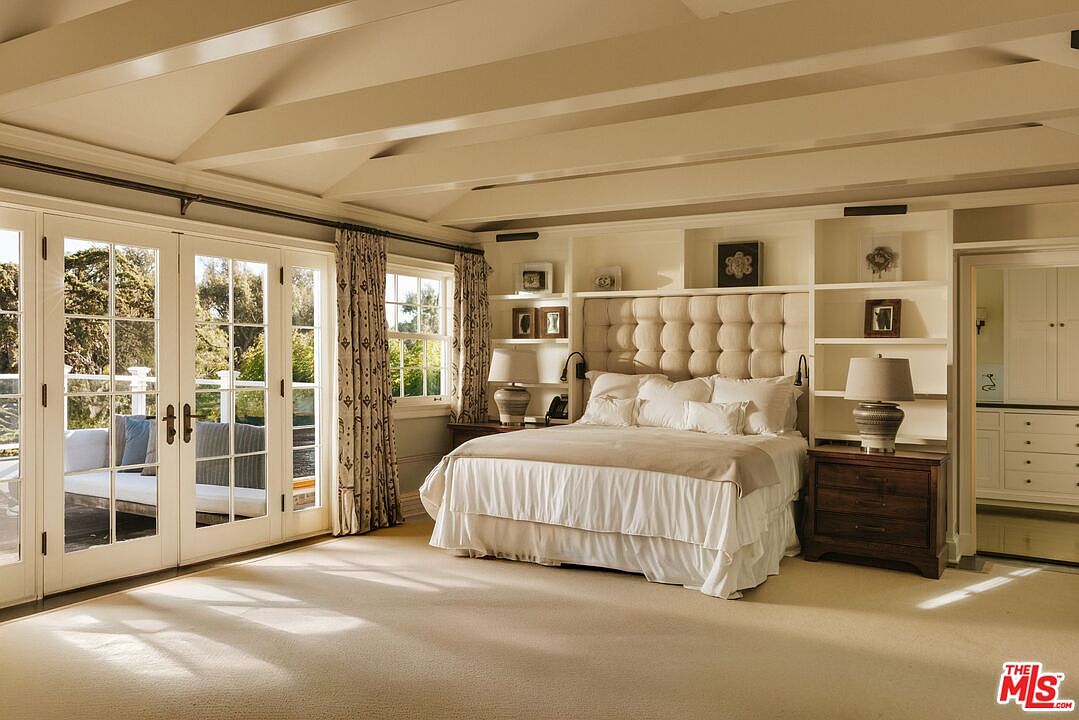 This elegant primary bedroom features a neutral color palette, a plush tufted headboard integrated into custom built-in shelving, and a vaulted ceiling with exposed beams. Large glass French doors open onto a private patio, flooding the room with natural light and creating a seamless indoor-outdoor connection. The space is anchored by a soft, light-colored carpet and warm, ambient lighting, evoking a serene and luxurious atmosphere.