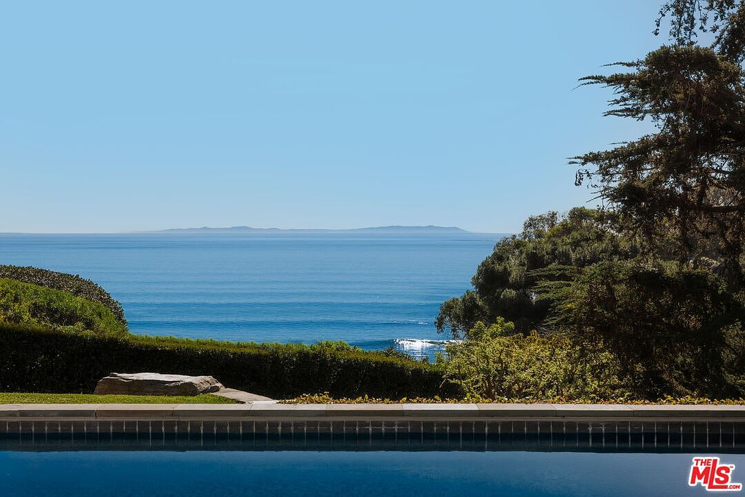 This serene image captures a stunning infinity-edge pool overlooking a vast, calm ocean with distant islands on the horizon. The foreground features a manicured hedge and a large, natural stone accent, creating a seamless transition between the private pool area and the expansive coastal landscape. The perspective is low and wide, emphasizing the tranquil atmosphere and the luxurious, unobstructed waterfront view.