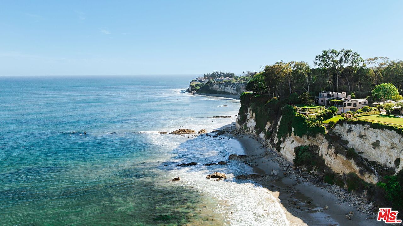 This stunning aerial perspective captures a luxurious coastal estate perched atop a dramatic cliffside overlooking the Pacific Ocean. The property features expansive green lawns and lush landscaping that seamlessly blend with the rugged natural coastline and sandy beach below. The wide-angle shot emphasizes the property's secluded, prime location and its breathtaking panoramic ocean views.