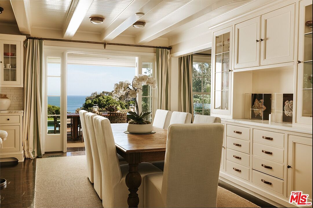 This elegant dining room features a warm wooden table surrounded by upholstered chairs, set against a backdrop of custom white built-in cabinetry. Large glass doors open directly to a scenic ocean view, seamlessly blending indoor dining with the outdoor landscape. The room is characterized by its bright, airy atmosphere, highlighted by a coffered ceiling and soft, neutral tones that create a sophisticated coastal aesthetic.
