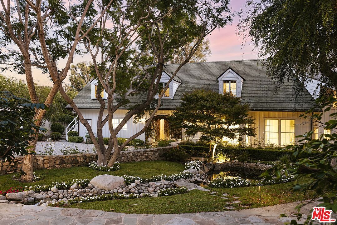 This charming, traditional-style home features a classic white board-and-batten facade with a steep, shingled roof and symmetrical dormer windows. The front yard is beautifully landscaped with a stone-lined pond, lush green lawn, and mature trees, creating a serene and inviting curb appeal. The warm glow from the interior lights and the soft evening sky enhance the picturesque, storybook quality of the property.