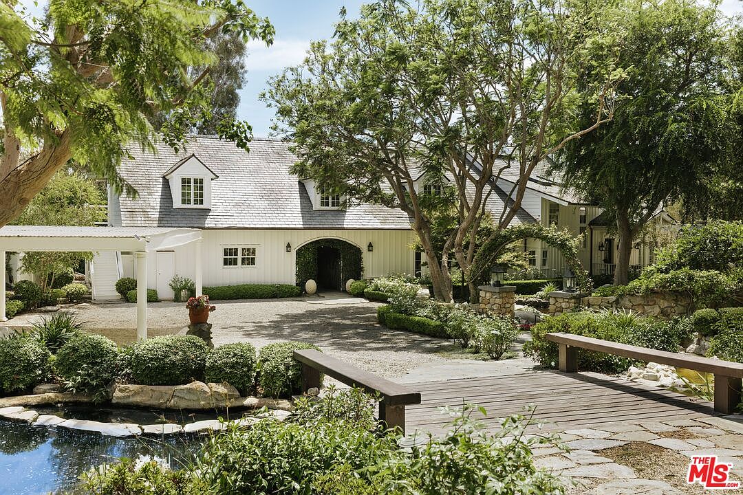 This charming, white-sided farmhouse features a classic gabled roof with dormer windows and a prominent arched entryway. The property is set back from a tranquil pond with a wooden footbridge in the foreground, surrounded by lush, mature trees and meticulously landscaped greenery. The gravel driveway leads up to the home, creating a serene and inviting atmosphere that exudes rustic elegance.