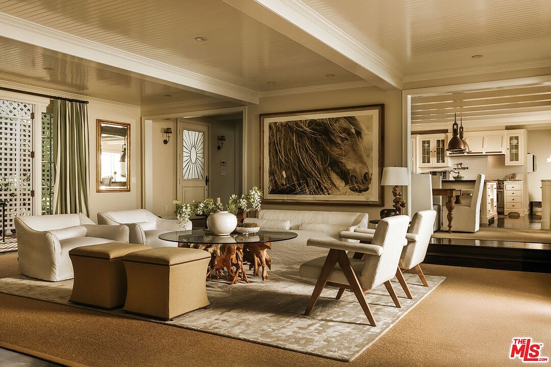This elegant living room features a sophisticated neutral color palette, anchored by a large, textured area rug and a striking coffee table with a glass top and natural wood base. A prominent horse portrait hangs above a plush white sofa, while mid-century modern armchairs and matching ottomans provide comfortable seating. The space flows seamlessly into an adjacent kitchen area, creating an open and inviting atmosphere with high-end finishes and architectural ceiling details.
