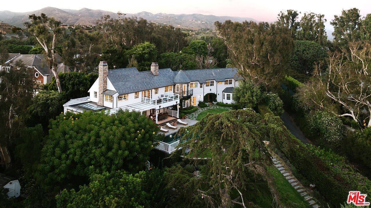 This high-angle aerial shot captures a sprawling, traditional-style estate nestled within a lush, tree-filled landscape. The white-painted home features a dark shingled roof, multiple chimneys, and a charming turret, complemented by a private swimming pool and a well-manicured lawn. The perspective highlights the property's expansive grounds and its serene, elevated position against a backdrop of rolling hills.