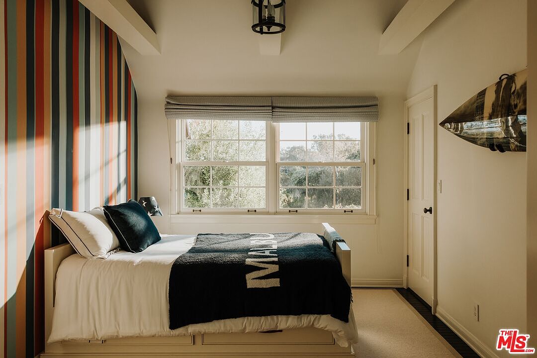 This cozy bedroom features a twin bed with a navy blue 'Malibu' throw blanket, set against a vibrant, multi-colored striped accent wall. A large window with a textured Roman shade allows natural light to fill the room, while a surfboard mounted on the wall adds a playful, coastal touch. The space is finished with neutral carpeting and a classic white door, creating a bright and inviting atmosphere.