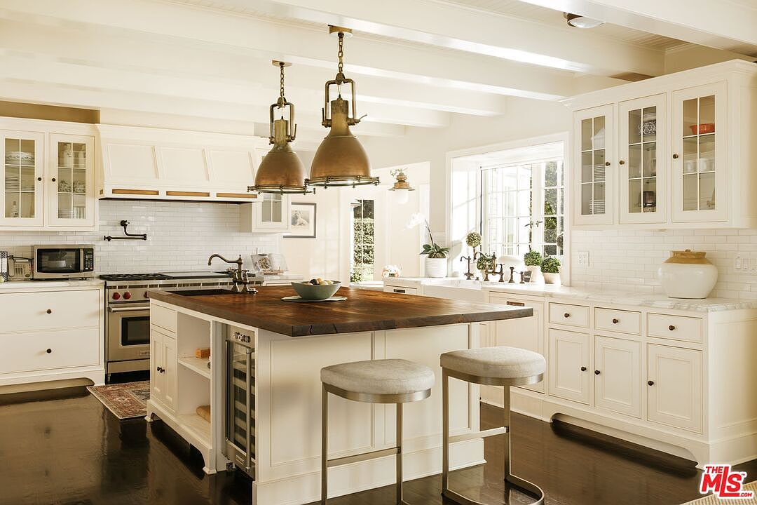 This bright and airy kitchen features a classic farmhouse aesthetic with white cabinetry, a large central island topped with a rich, dark wood butcher block, and elegant pendant lighting. The space is equipped with high-end stainless steel appliances, a subway tile backsplash, and marble countertops, creating a warm and inviting atmosphere. The perspective is a wide-angle shot from the kitchen floor, showcasing the seamless flow between the cooking area and the breakfast bar seating.