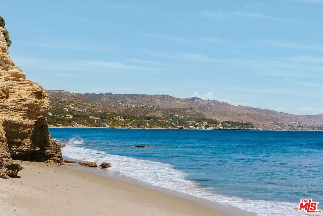 This scenic coastal view captures a sandy beach meeting the deep blue Pacific Ocean, framed by rugged, light-colored sandstone cliffs on the left. In the distance, a rolling coastline features hillside residential properties nestled against a backdrop of arid, mountainous terrain under a clear, bright sky. The perspective is a wide, eye-level shot that emphasizes the natural beauty and serene atmosphere of the beachfront location.