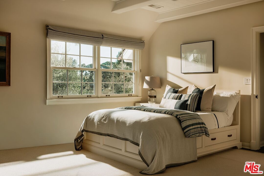 This cozy and well-lit bedroom features a neutral color palette with a comfortable bed dressed in layered linens and striped accent pillows. A large window with a fabric roman shade allows natural light to fill the room, highlighting the clean lines of the white bed frame with built-in storage drawers. The space is accented by a bedside lamp and a framed piece of art, creating a serene and inviting atmosphere.