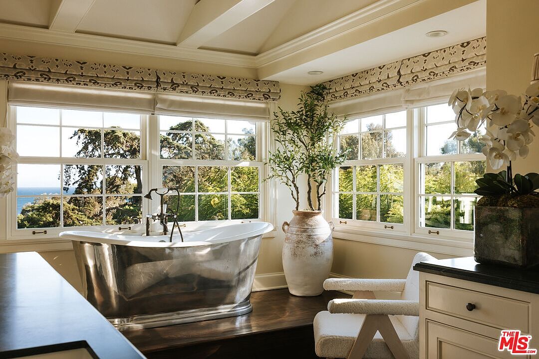 This luxurious primary bathroom features a striking metallic soaking tub positioned in front of large windows that offer a serene view of the ocean and lush greenery. The space is accented by a large, rustic ceramic floor vase with a tall plant and a comfortable, textured armchair, creating a spa-like atmosphere. The combination of dark wood flooring, neutral walls, and elegant window treatments provides a sophisticated and tranquil aesthetic.