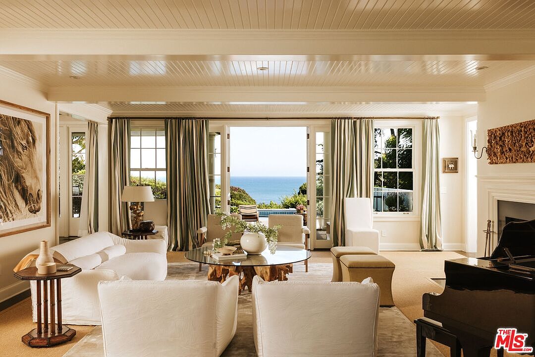 This elegant, light-filled living room features a sophisticated coastal aesthetic with plush white seating, a unique driftwood-base coffee table, and a grand piano. Large glass doors open to a stunning, unobstructed view of the ocean and a swimming pool, seamlessly blending indoor comfort with the natural beauty of the coastline. The room is accented by warm, neutral tones, a large horse-themed artwork, and classic architectural details like beadboard ceilings.