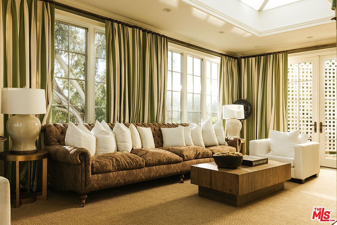 This elegant living room features a large, tufted brown patterned sofa adorned with numerous white throw pillows, centered around a minimalist wooden coffee table. The space is framed by floor-to-ceiling windows draped in sophisticated green and white striped curtains, allowing ample natural light to fill the room. A white armchair and matching table lamps add a bright, refined touch to the traditional and cozy atmosphere.