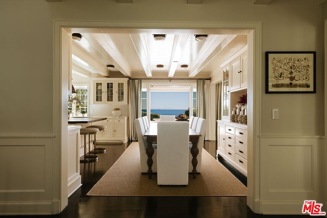 This elegant dining room features a long wooden table surrounded by white upholstered chairs, set against a backdrop of a stunning ocean view through large glass doors. The room is framed by a classic architectural archway, highlighting a coffered ceiling with recessed lighting and a built-in white cabinetry unit on the right. The space exudes a sophisticated, coastal-inspired aesthetic with a neutral color palette and a textured area rug.