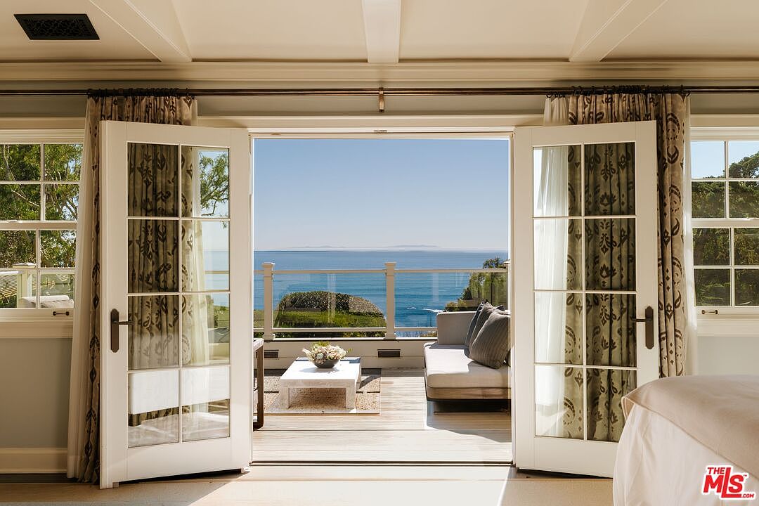 This view from inside a primary bedroom showcases elegant French doors opening onto a private balcony with a stunning, unobstructed ocean view. The balcony is furnished with a comfortable outdoor sofa and a modern coffee table, creating a seamless transition between indoor luxury and coastal living. The room features classic architectural details, including crown molding and patterned curtains, framing the picturesque horizon.