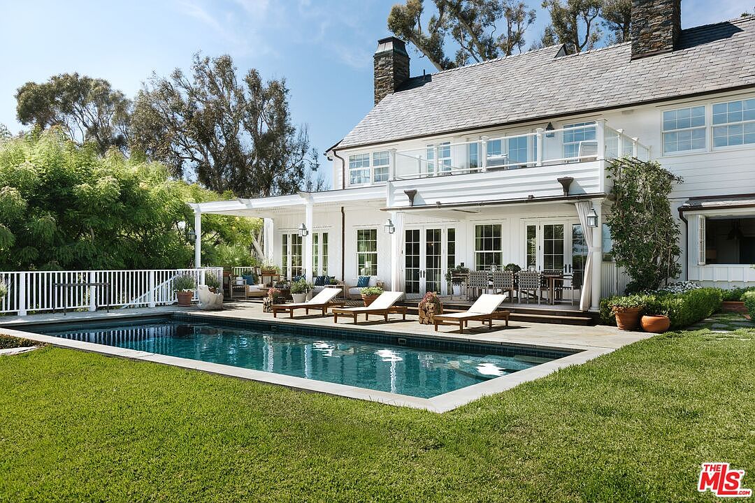 This stunning rear exterior view showcases a classic, multi-story white home featuring a spacious covered patio, a second-story balcony with glass railings, and a pristine rectangular swimming pool. The manicured lawn leads directly to the pool deck, which is furnished with lounge chairs, creating an inviting and luxurious outdoor living space. The architectural style is elegant and traditional, perfectly complemented by the lush surrounding trees and clear blue sky.