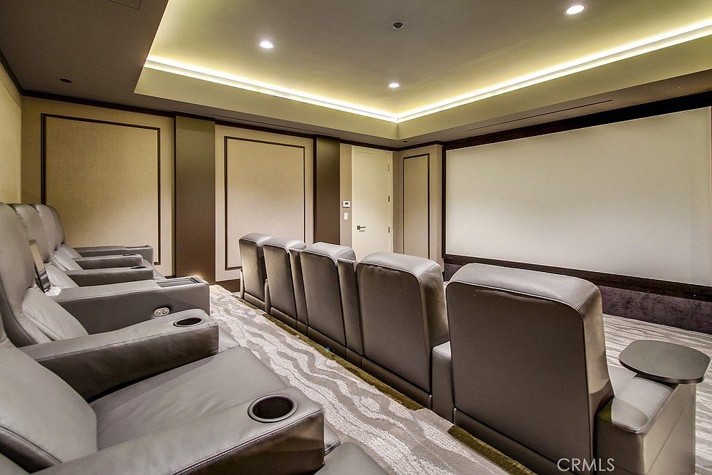 This dedicated home theater room features multiple rows of plush, grey leather reclining seats arranged for optimal viewing of a large projection screen. The space is designed with sophisticated recessed cove lighting in the ceiling and paneled wall accents, creating a professional cinematic atmosphere. The room is carpeted with a patterned runner, and the overall aesthetic is modern, luxurious, and focused on entertainment.