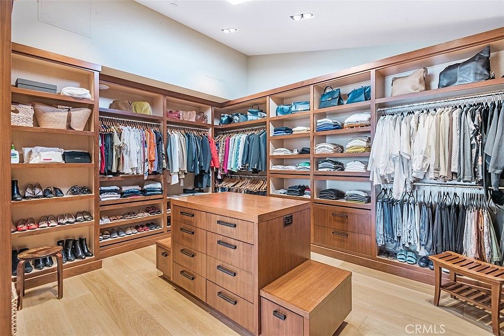 This expansive walk-in closet features custom wood cabinetry with extensive hanging space, open shelving, and a central island with multiple drawers. The room is finished with light wood flooring and recessed lighting, creating a bright and organized atmosphere. A small wooden bench is positioned near the entrance, adding a functional touch to this well-appointed storage space.