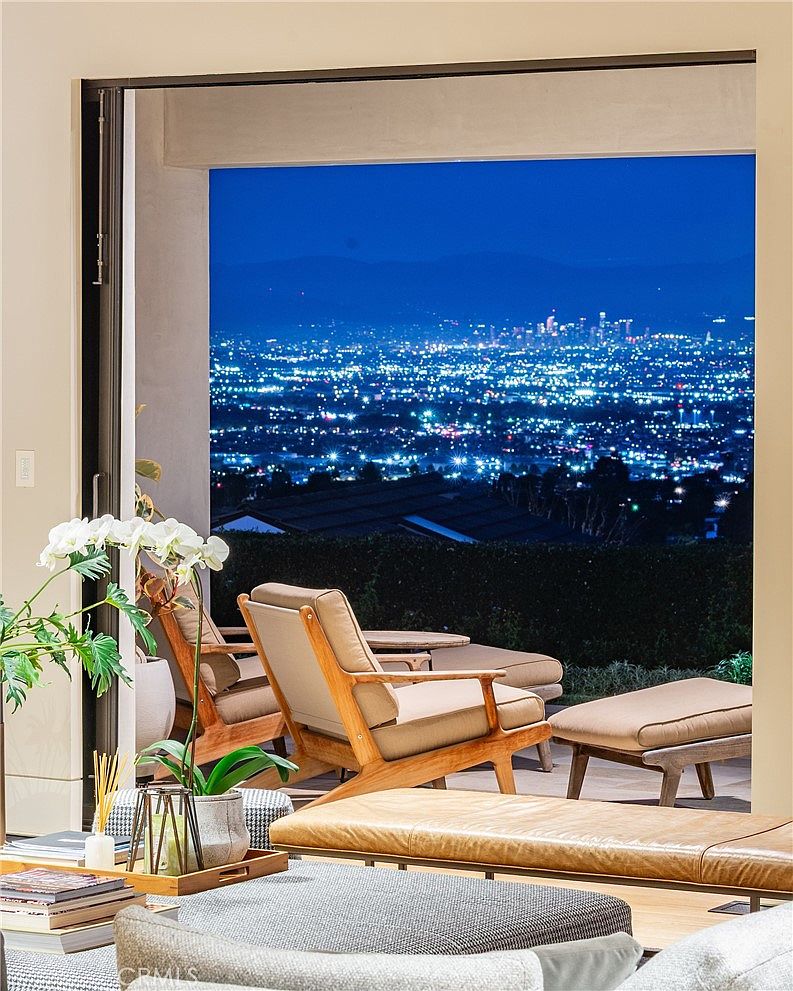 This interior living space features a seamless transition to an outdoor patio through large, open glass doors that frame a stunning panoramic view of a city skyline at night. The foreground includes comfortable light wood lounge chairs with neutral cushions and a low-profile leather bench, creating a sophisticated and relaxing atmosphere. The composition emphasizes the connection between the refined indoor living area and the expansive, illuminated urban landscape beyond.