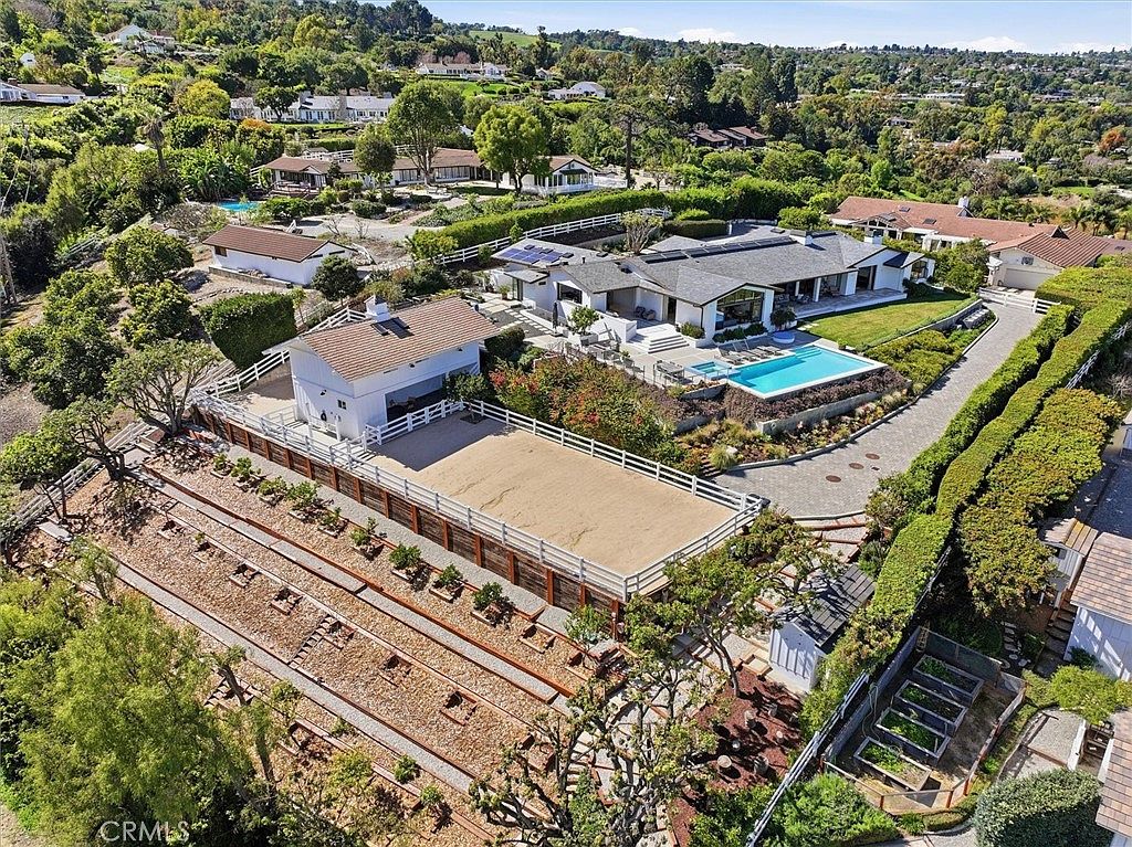 This high-angle aerial view captures a sprawling estate featuring a modern, single-story residence with a swimming pool and a dedicated equestrian riding arena. The property is surrounded by lush greenery, mature trees, and terraced landscaping, showcasing a blend of luxury living and rural functionality. The expansive grounds include a paved driveway, a detached barn-style structure, and a well-maintained outdoor space perfect for recreation and relaxation.