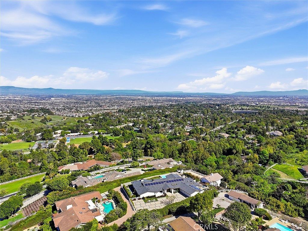 This high-angle aerial perspective captures a sprawling residential property nestled within a lush, tree-filled landscape. The primary residence features a dark-tiled roof with integrated solar panels, a paved motor court, and a private swimming pool, all set against a vast, panoramic backdrop of a suburban valley and distant mountains. The composition emphasizes the home's secluded, elevated position and its expansive outdoor living potential.