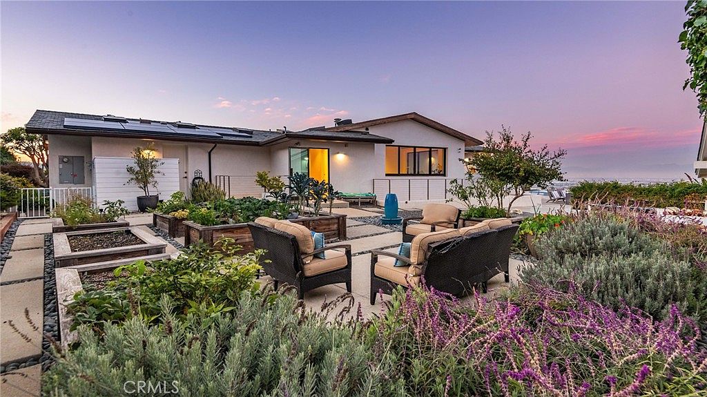 This serene outdoor space features a landscaped garden with raised wooden planter boxes and a paved patio area furnished with comfortable outdoor seating. The home's exterior is finished in a light, neutral tone with solar panels visible on the roof, set against a soft, twilight sky. The perspective captures a peaceful, inviting backyard retreat that seamlessly blends architectural design with natural beauty.