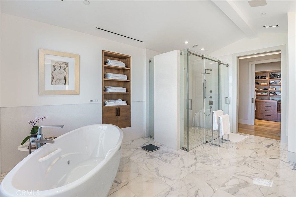 This bright and airy primary bathroom features a freestanding soaking tub as the focal point, set against a backdrop of white walls and patterned stone flooring. A glass-enclosed walk-in shower sits adjacent to a built-in shelving unit, while a doorway leads into a spacious walk-in closet with custom wood cabinetry. The space is illuminated by recessed lighting, creating a clean, luxurious, and spa-like atmosphere.