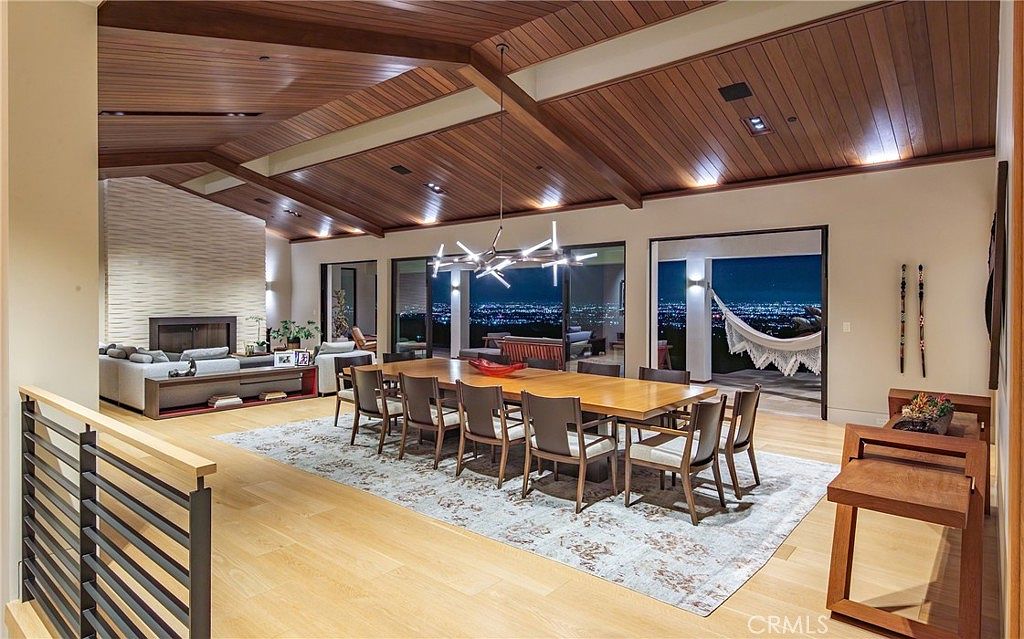 This expansive open-concept living and dining area features a dramatic vaulted wood-paneled ceiling and light-toned wood flooring. A long dining table serves as the centerpiece, complemented by a modern geometric chandelier, while a cozy living area with a textured stone chimney breast sits to the left. Large glass sliding doors open to a balcony, revealing a stunning panoramic view of the city lights at night.