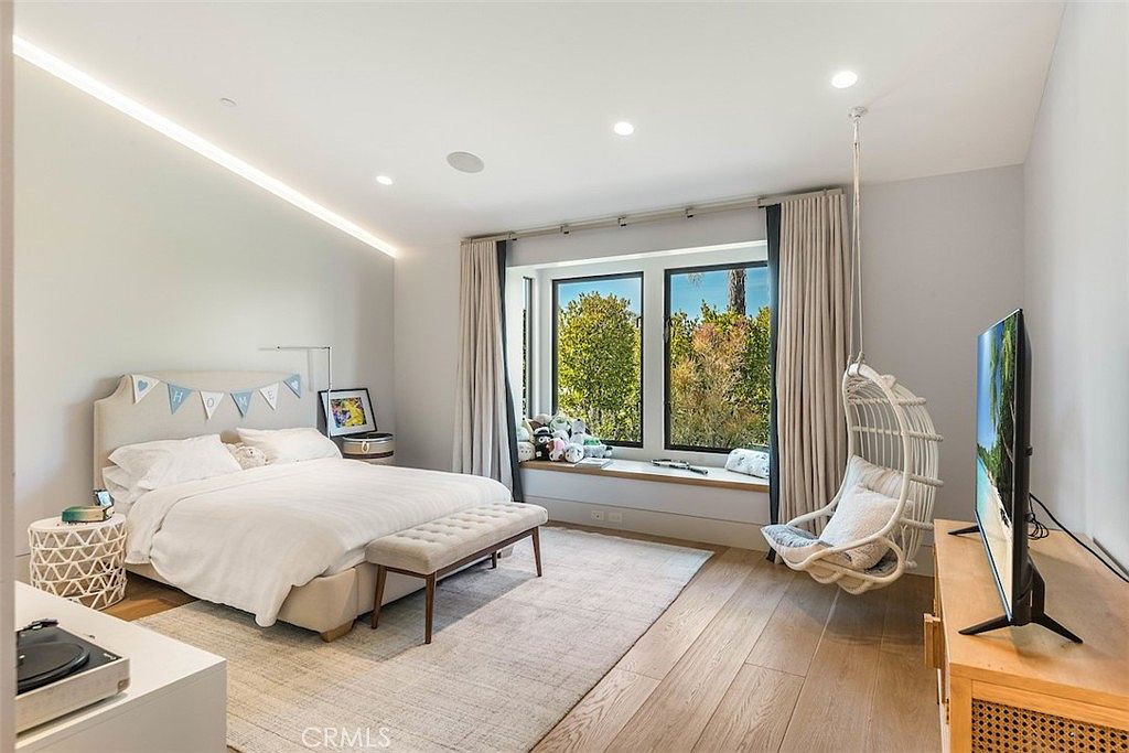 This bright and airy bedroom features light wood flooring, a neutral color palette, and a large window with a built-in bench seat overlooking greenery. The room is furnished with a queen-sized bed, a tufted bench at the foot, and a hanging wicker chair, creating a relaxed and inviting atmosphere. Recessed lighting and a minimalist aesthetic contribute to the clean, modern feel of the space.
