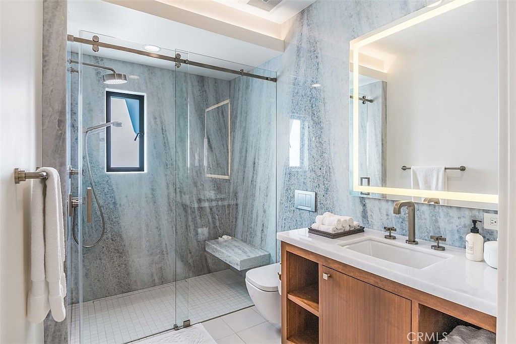 This modern bathroom features striking blue-toned stone wall cladding that extends into the walk-in shower area. The space includes a floating wood vanity with a white countertop, a sleek illuminated mirror, and a glass-enclosed shower with a built-in stone bench. The perspective captures the entire vanity and shower layout, highlighting the contemporary design and high-end finishes.