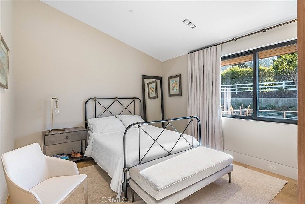 This bright and airy bedroom features a metal-framed bed centered against a neutral-toned wall, complemented by a matching nightstand and a white upholstered bench at the foot of the bed. A large window with light-colored curtains allows natural light to fill the space, highlighting the light wood flooring and the minimalist decor. The room is accented by a modern white armchair in the foreground and two framed pictures hanging above the bed, creating a clean and inviting atmosphere.