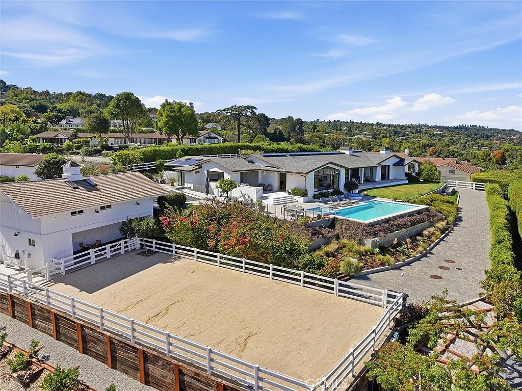 This high-angle aerial perspective showcases a sprawling, single-story ranch-style estate featuring a white exterior and a multi-gabled roofline. The property includes a large rectangular swimming pool, a dedicated equestrian riding arena with white fencing, and a separate detached structure. The expansive grounds are surrounded by lush greenery and rolling hills, creating a serene and private atmosphere.